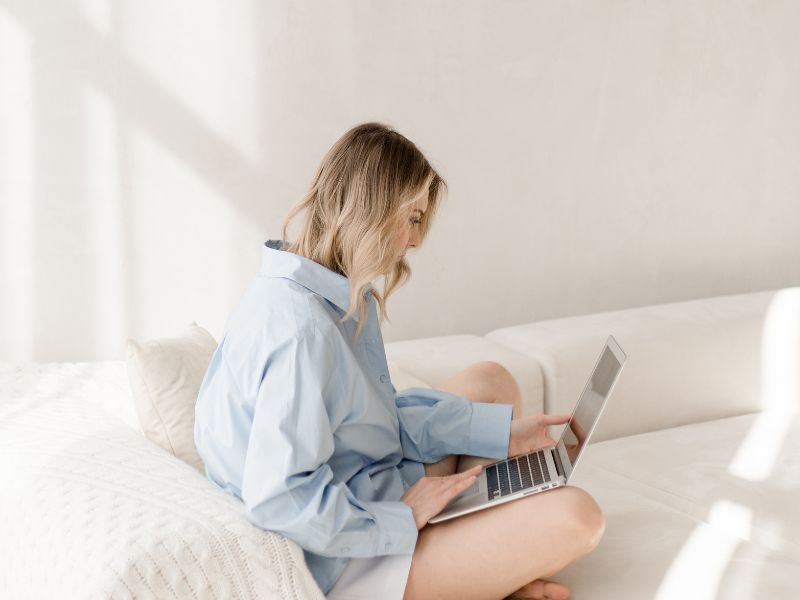 Lohnt sich Bloggen noch in 2025? 3 Frau in blauer Bluse sitzt auf Sofa in beige und arbeitet remote am Laptop von zuhause aus. Bloggen lohnt sich auch 2025 noch