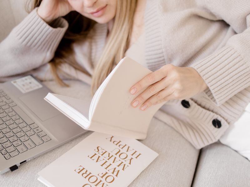 Blonde Frau liegt mit Laptop und Motivationsbuch auf Sofa und will sich nebenberuflich selbstständig machen.